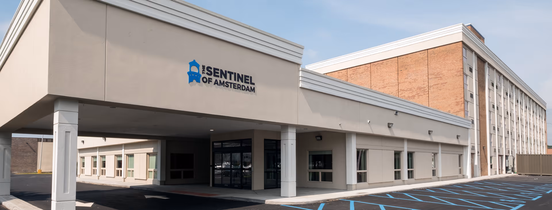 Exterior view of The Sentinel of Amsterdam senior living facility showing the main entrance with a covered drop-off area, beige and brick building facade, and an empty parking lot with blue handicap parking markings.