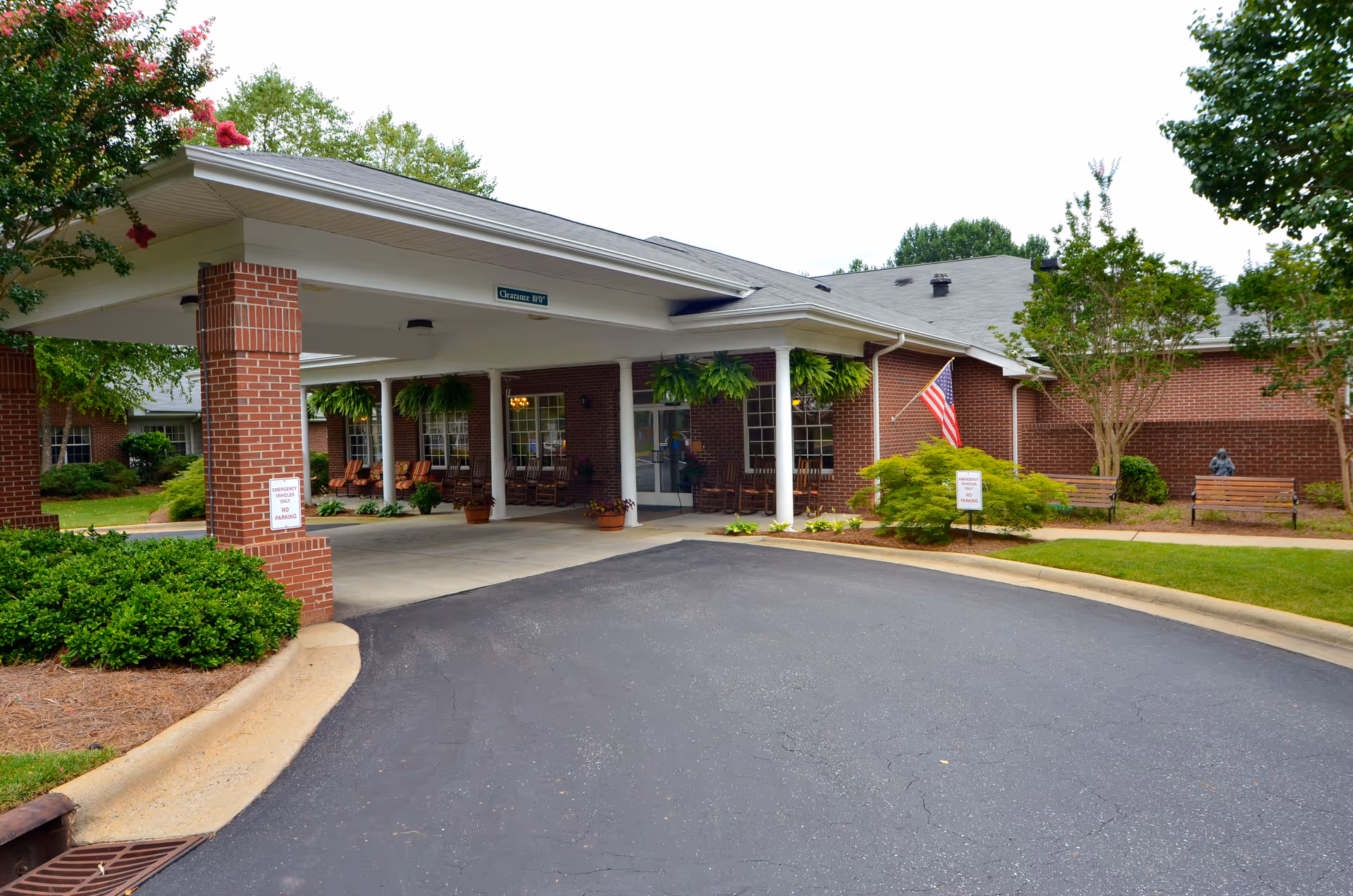 Entrance of a brick building with a covered driveway supported by brick and white columns. There are hanging plants under the covered area, several chairs, an American flag, and landscaped greenery around the entrance.