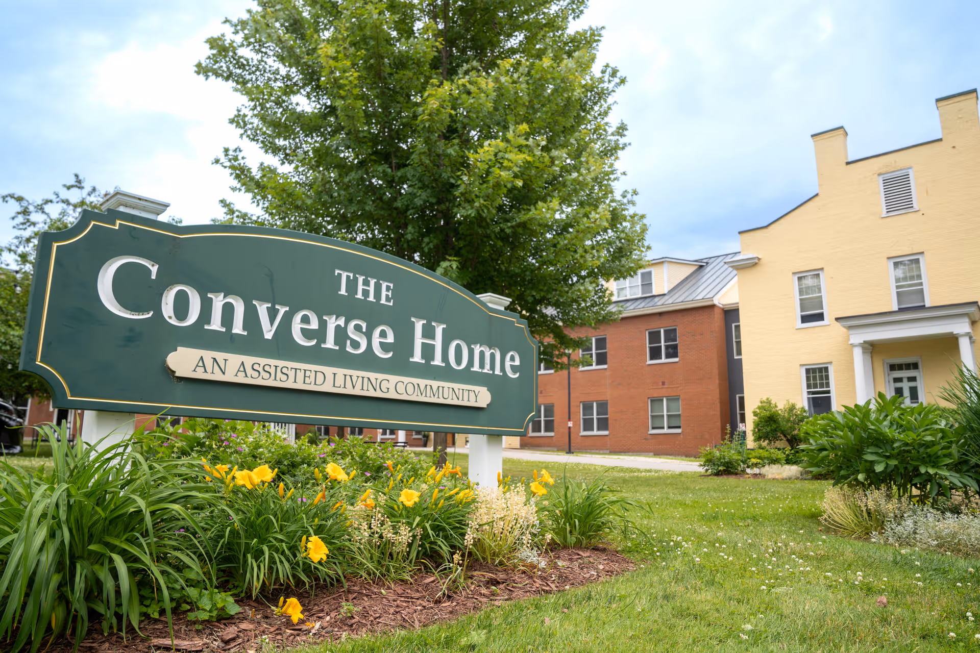 Outdoor view of The Converse Home assisted living community sign surrounded by flowers and greenery, with residential buildings and a tree in the background under a partly cloudy sky.