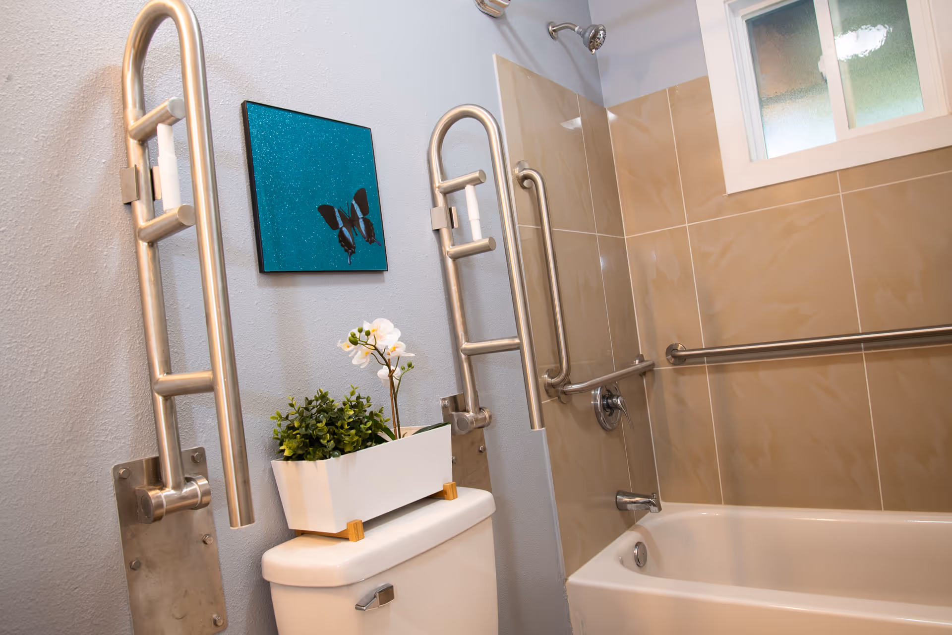 A bathroom with beige tiled walls and a white bathtub equipped with multiple stainless steel grab bars for accessibility. There is a white toilet with a white planter holding green and white artificial plants on top. A small window with frosted glass is above the bathtub, and a blue wall decoration featuring a butterfly is mounted on the wall.