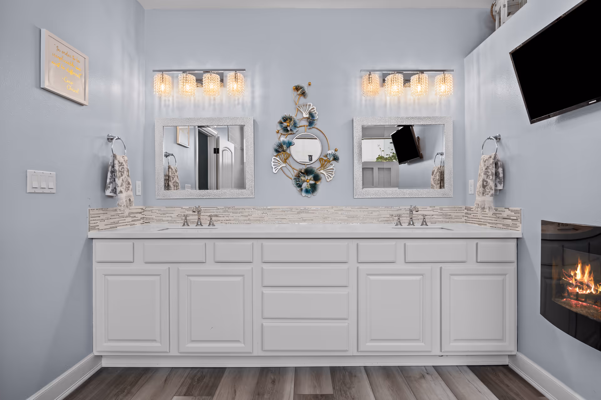 Dual-sink bathroom vanity with two mirrors, decorative wall art, modern light fixtures, and a wall-mounted TV and fireplace.