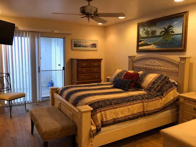 A cozy bedroom with a large bed featuring a patterned bedspread and multiple pillows. The room includes a wooden dresser, a small bench at the foot of the bed, a chair, and a ceiling fan. There is a sliding glass door with vertical blinds leading to an outdoor area. The walls are decorated with framed artwork, including a painting of palm trees above the bed.