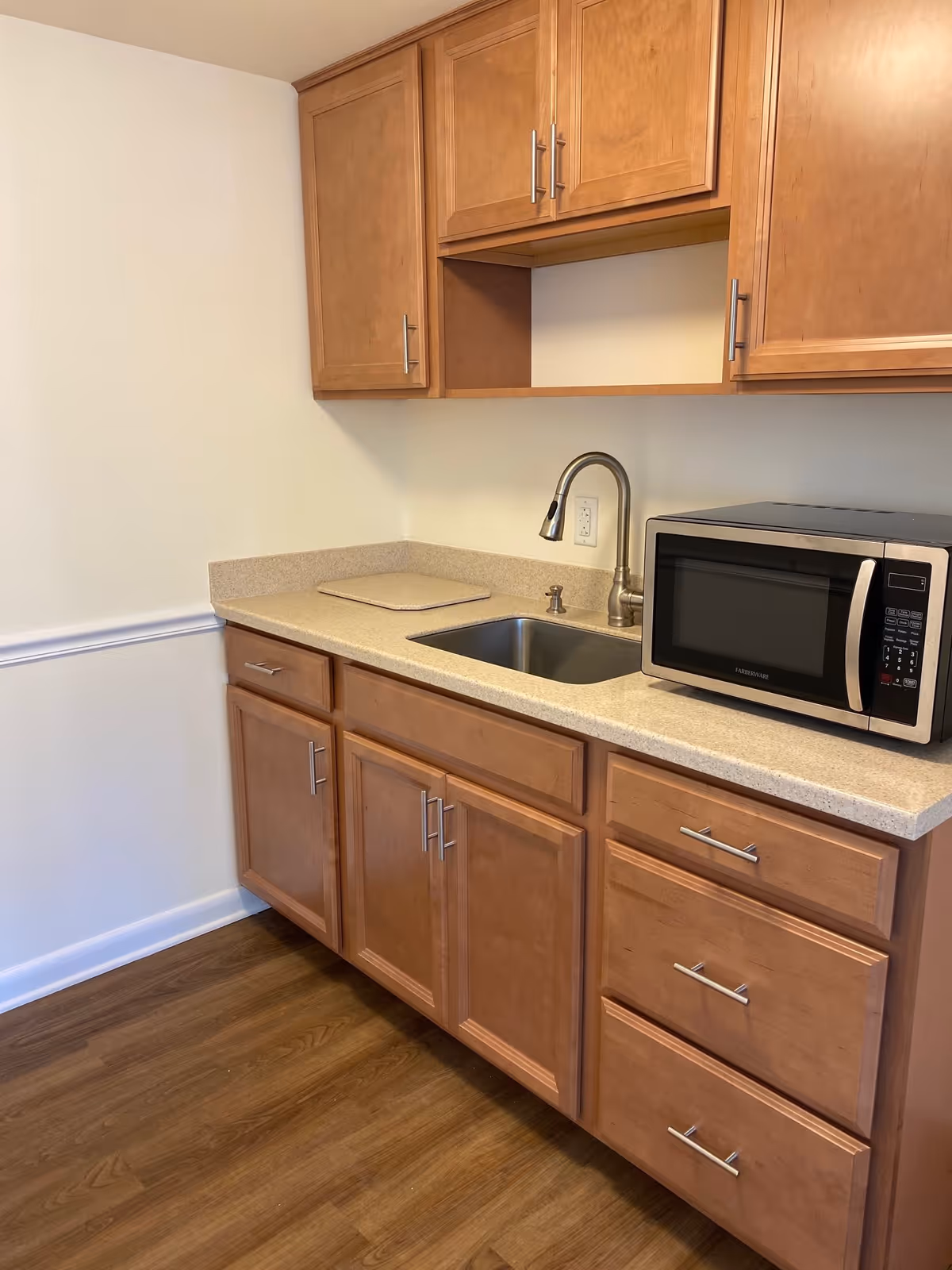 A kitchenette area with wooden cabinets, a countertop with a built-in sink and faucet, and a microwave oven placed on the right side of the counter. The floor is wooden, and the walls are painted white with a chair rail molding.