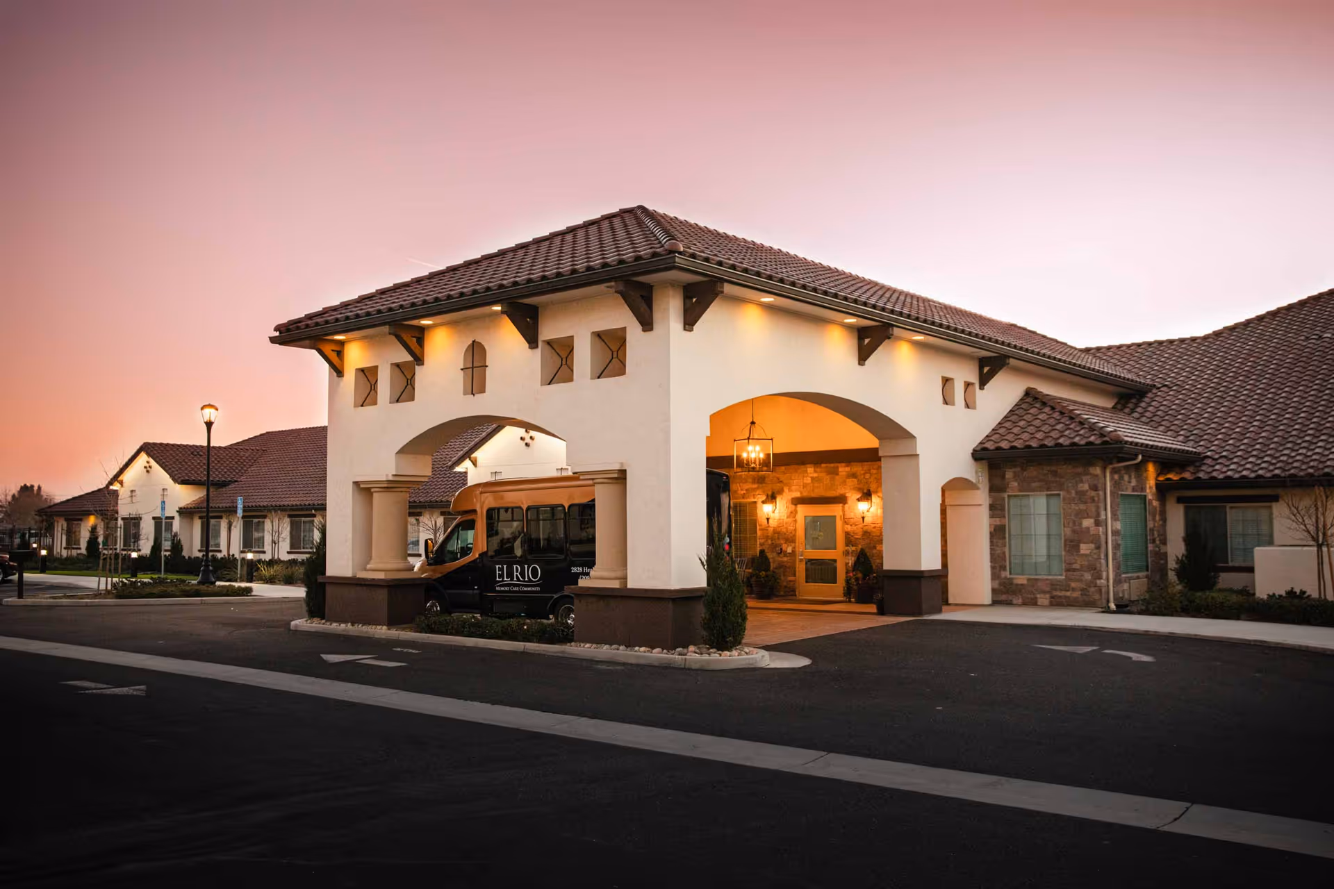 Exterior view of El Rio Memory Care Community building at dusk, featuring a covered entrance with arches, warm lighting, and a parked shuttle van with the facility's name on it.