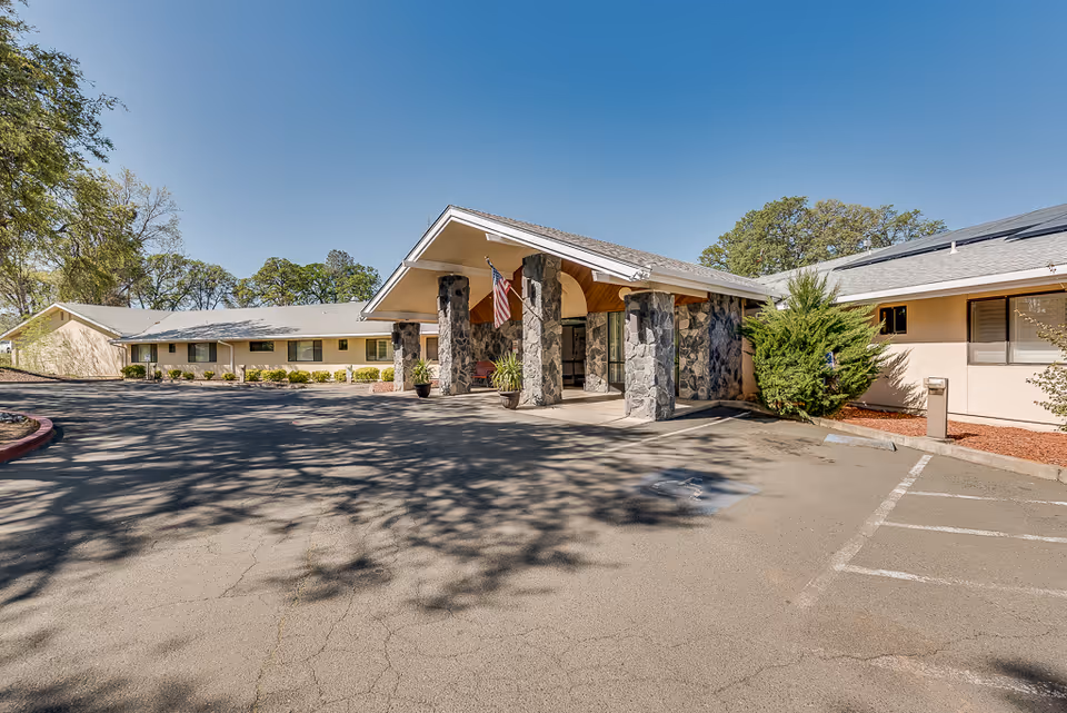 Exterior view of Meadowood Nursing Center showing a single-story building with a covered entrance supported by stone pillars, an American flag hanging, and a parking area in front under a clear blue sky.
