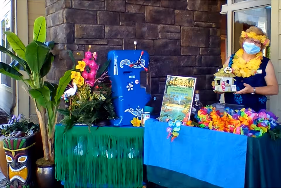 A woman wearing a face mask and a yellow flower lei stands behind a table decorated with colorful leis, tropical flowers, and a blue ukulele. There is a sign that reads 'Hawaii' and a tiki statue plant pot on the left side. The setting appears to be an outdoor or semi-outdoor area with a stone wall background.