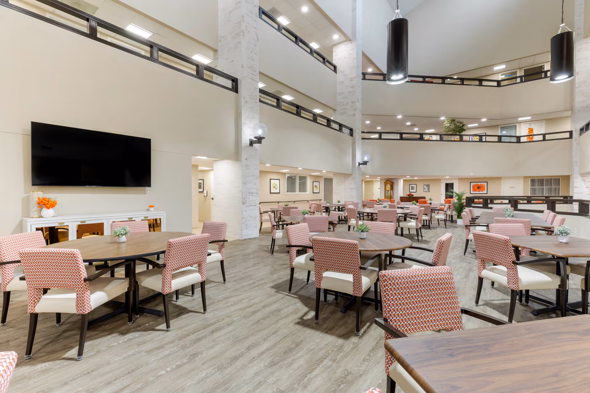 A spacious, well-lit dining area in a senior living facility with multiple round wooden tables surrounded by cushioned chairs with patterned upholstery. The room features high ceilings with modern pendant lights, a large flat-screen TV mounted on the wall, and decorative plants on the tables. The area is open to an upper level with railings and has a clean, contemporary design.