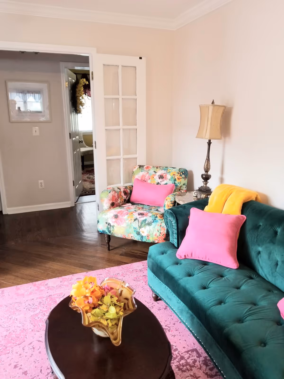 A cozy living room with a green velvet tufted sofa adorned with pink and yellow pillows, a floral patterned armchair with a pink pillow, a wooden coffee table with a decorative glass bowl containing yellow and orange flowers, a pink area rug, and a tall table lamp with a beige shade. A white door with glass panels is open, revealing another room with a window and a wreath on the door.