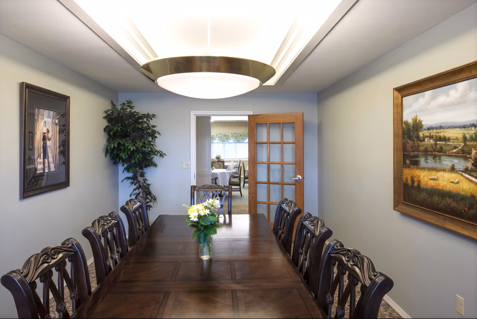 A formal dining room with a long wooden table surrounded by ornate wooden chairs. A vase with white and yellow flowers is placed in the center of the table. The room has light gray walls adorned with framed paintings and a large ceiling light fixture. A wooden door with glass panels is open, revealing another dining area with tables and chairs in the background.