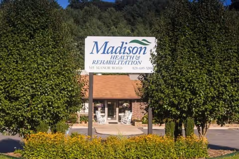 Sign reading Madison Health and Rehabilitation near entrance, framed by trimmed trees.