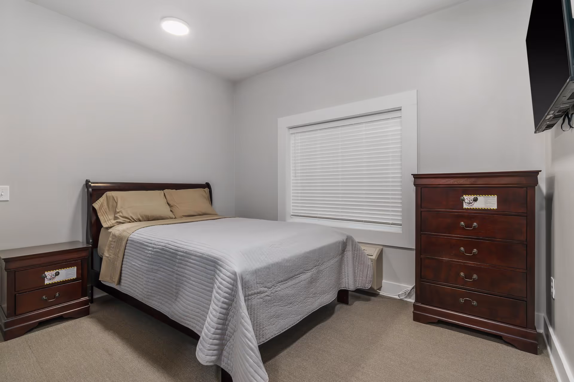 Neat bedroom with a made double bed, wooden nightstand and tall dresser, a window with blinds and a wall-mounted TV.