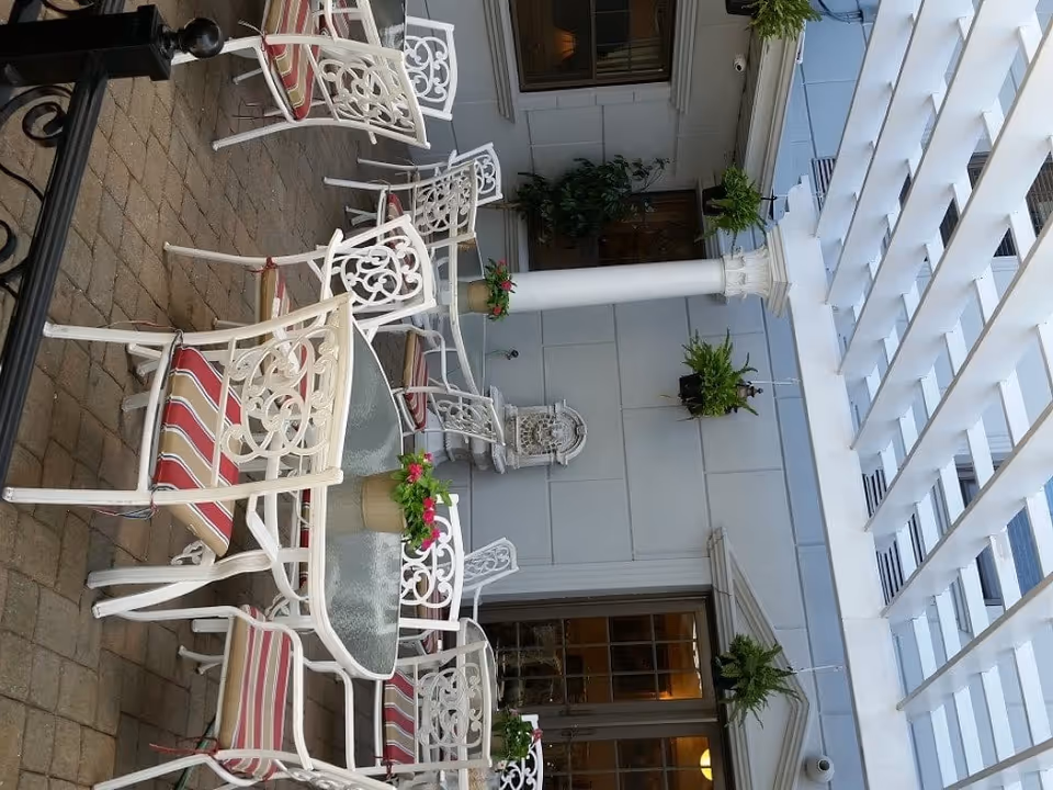 Outdoor patio area with white metal chairs and tables featuring red and white striped cushions. The patio has a brick floor, white columns, hanging green plants, and a white pergola overhead. There are windows and a door leading inside the building in the background.