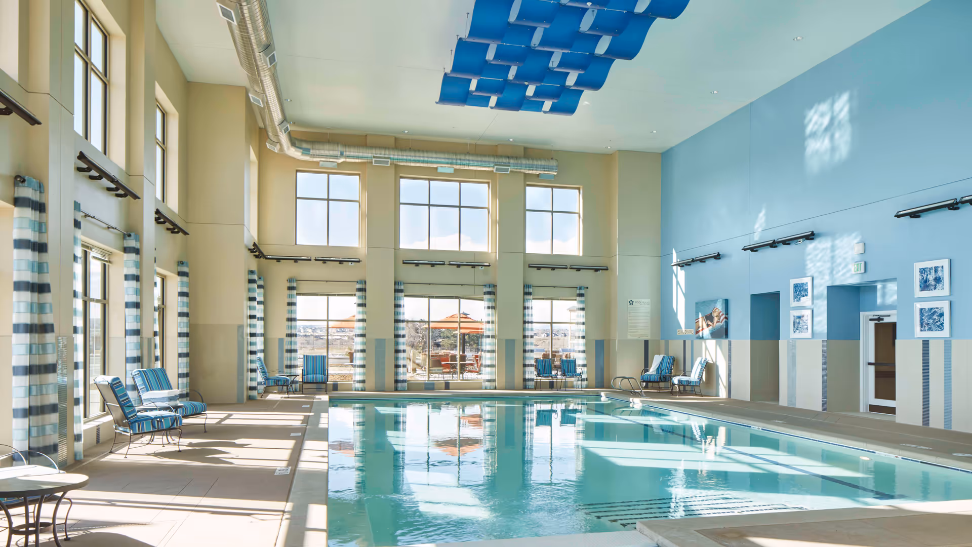 Indoor swimming pool area with large windows letting in natural light, blue and white striped curtains, several lounge chairs with blue cushions along the sides, and blue ceiling decorations above the pool.