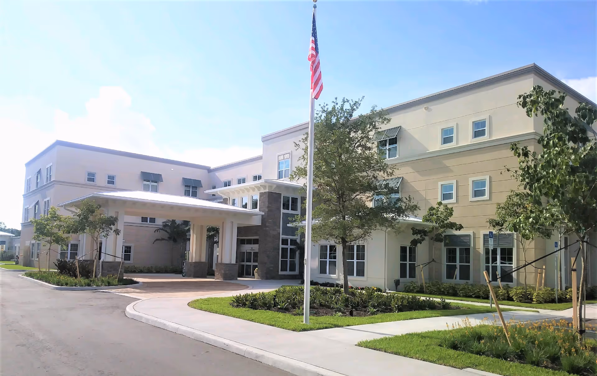 Exterior view of a modern three-story senior living facility building with a covered entrance, landscaped greenery, and an American flag on a flagpole in front.
