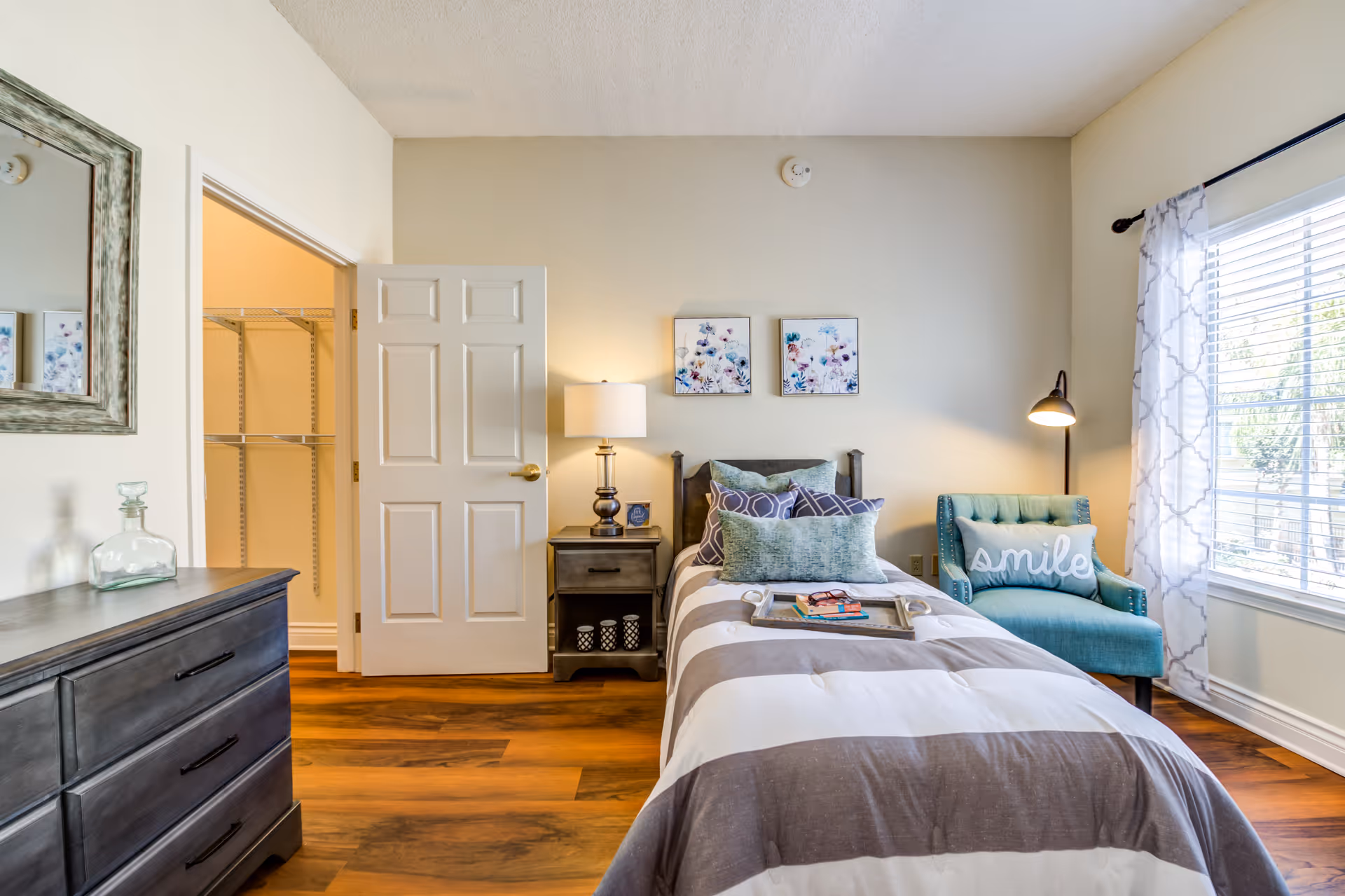 A cozy bedroom with a single bed covered in a striped gray and white comforter and multiple pillows. Next to the bed is a nightstand with a lamp and decorative items. A teal armchair with a pillow that says 'smile' is placed near a window with white patterned curtains. A dresser with a glass bottle and a large mirror above it is on the left side. The door to a walk-in closet is open, revealing shelving inside. The room has wooden flooring and light-colored walls.