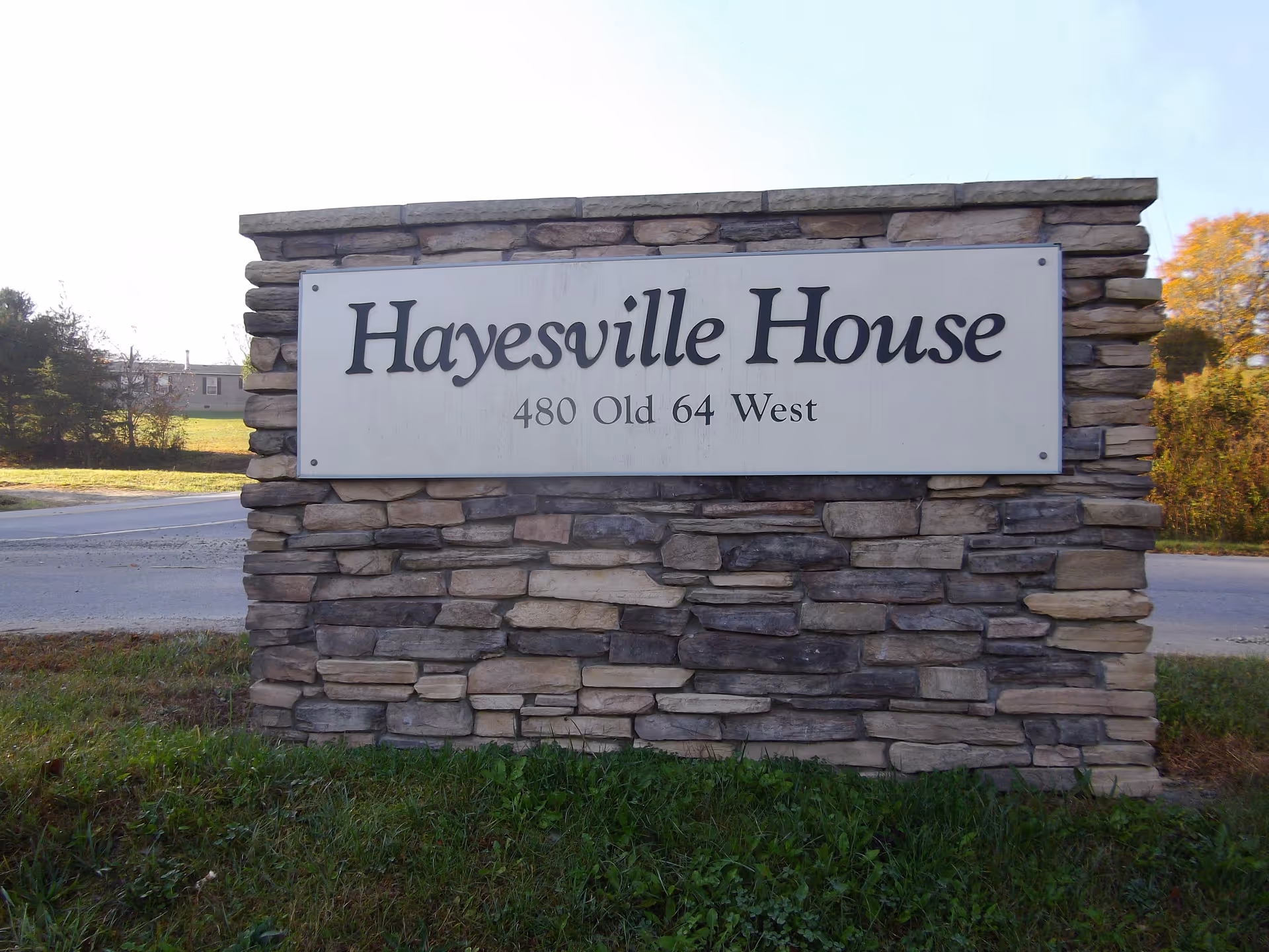 A stone sign with a white rectangular plaque that reads 'Hayesville House 480 Old 64 West' situated on a grassy area near a road with trees and buildings in the background.