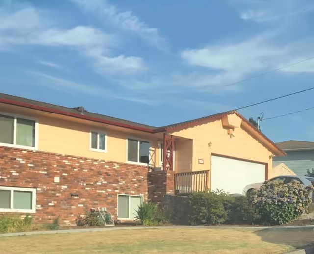 Exterior view of a single-story residential building with a brick and yellow siding facade, a garage, a small porch with railing, and a driveway with a parked car. There are bushes and flowers near the driveway and a clear blue sky with some clouds above.