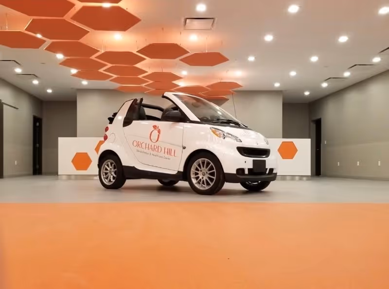 A small white Orchard Hill–branded Smart car parked in a modern indoor lobby with orange hexagonal ceiling panels and accents.