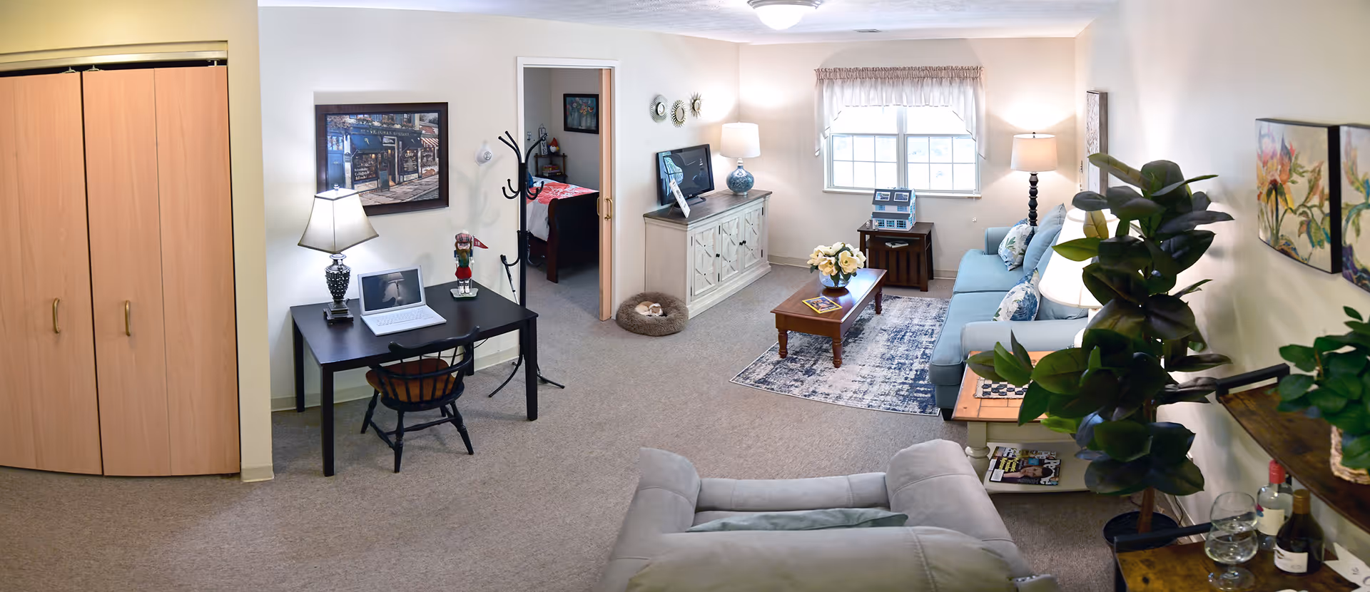 A cozy living room area with a light blue sofa, a wooden coffee table with flowers, a patterned rug, and a large window with curtains. To the left, there is a black desk with a laptop, lamp, and chair next to a wooden closet. A doorway leads to a bedroom with a bed visible. The room is decorated with plants, paintings, and lamps, creating a warm and inviting atmosphere.