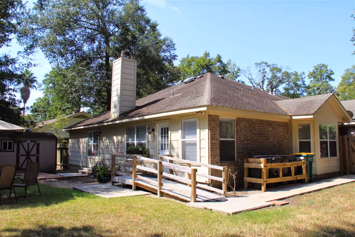 Single-story brick and siding cottage with a wooden accessibility ramp and small backyard patio under trees.