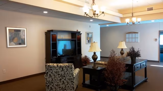 A cozy living room area in a senior living facility featuring a patterned armchair, a dark wood entertainment center with a TV, two table lamps on side tables, decorative plants, and framed artwork on beige walls under ceiling lights.