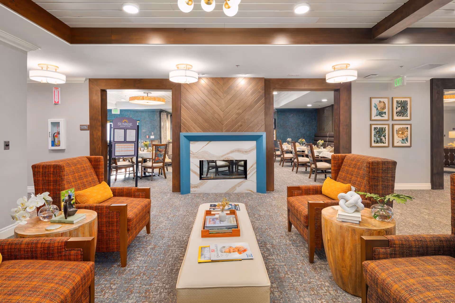 A cozy senior living facility lounge area with four orange plaid armchairs arranged around a central beige ottoman. The ottoman has magazines and a small vase on it. There are two wooden side tables with decorative items and flowers. In the background, there is a modern fireplace with a blue frame and wood paneling above it. Beyond the fireplace, a dining area with tables and chairs is visible. The walls are decorated with framed botanical prints and the ceiling has recessed lighting and modern light fixtures.