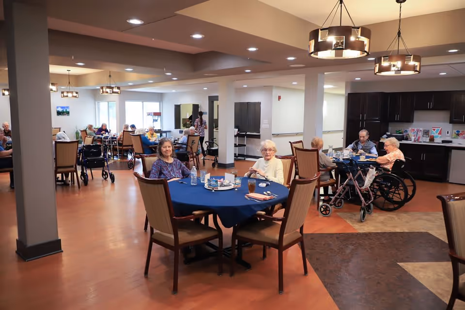 Seniors seated at round tables in a bright communal dining room with wood floors and pendant lights.