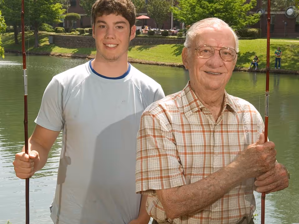 An elderly man and a young man standing side by side outdoors near a pond, each holding a fishing rod, with green grass and trees in the background.