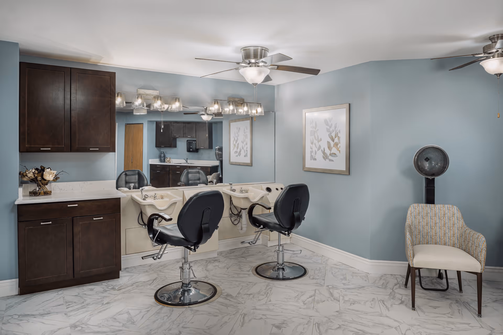 Interior view of a senior living facility's hair salon area with two black salon chairs in front of wash basins, dark wood cabinets, a large mirror, a ceiling fan with light, a framed botanical artwork on a blue wall, a vintage hair dryer, and a cushioned chair.