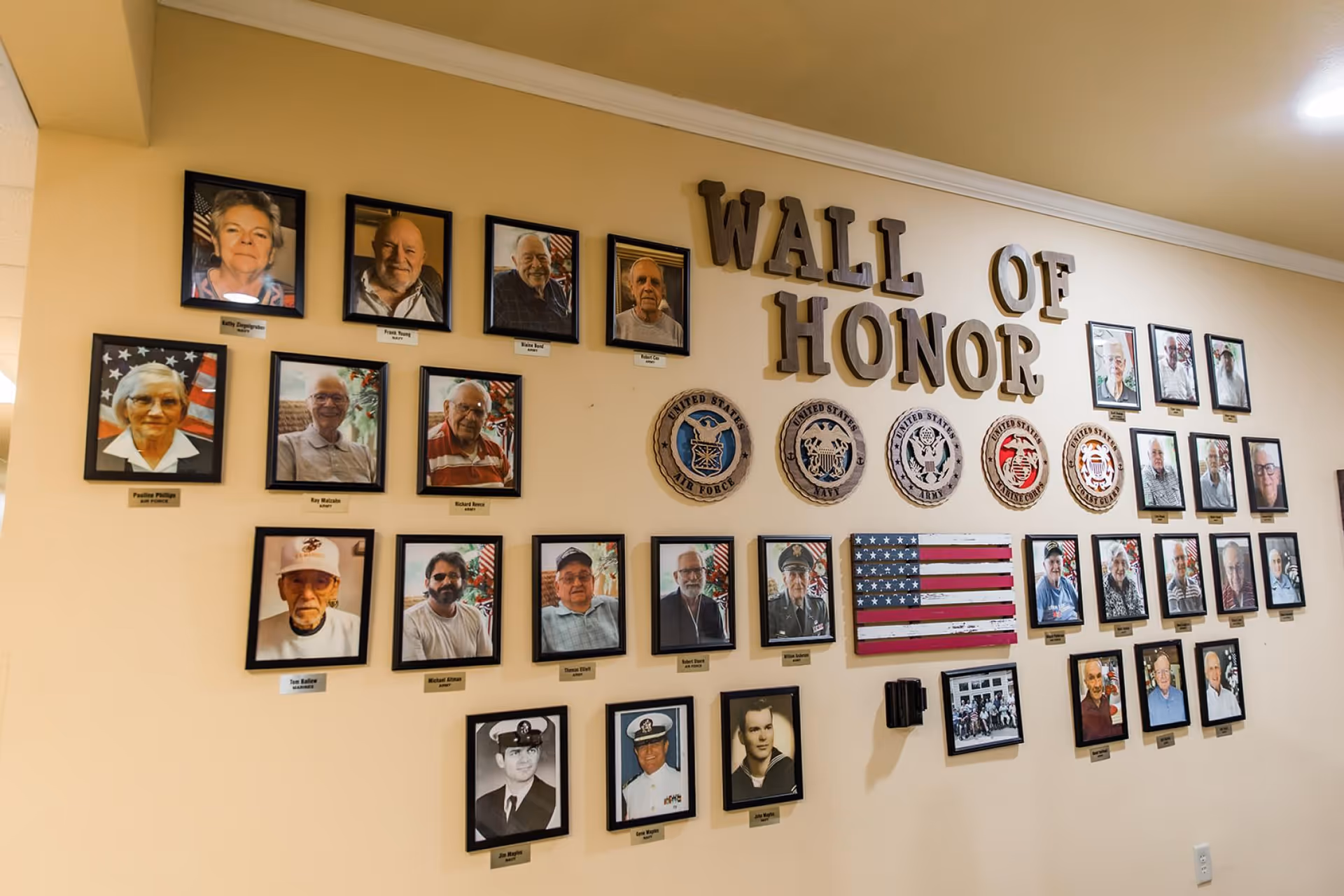 An interior 'Wall of Honor' display with framed portraits of residents, military emblems, and a small American flag on a beige wall.