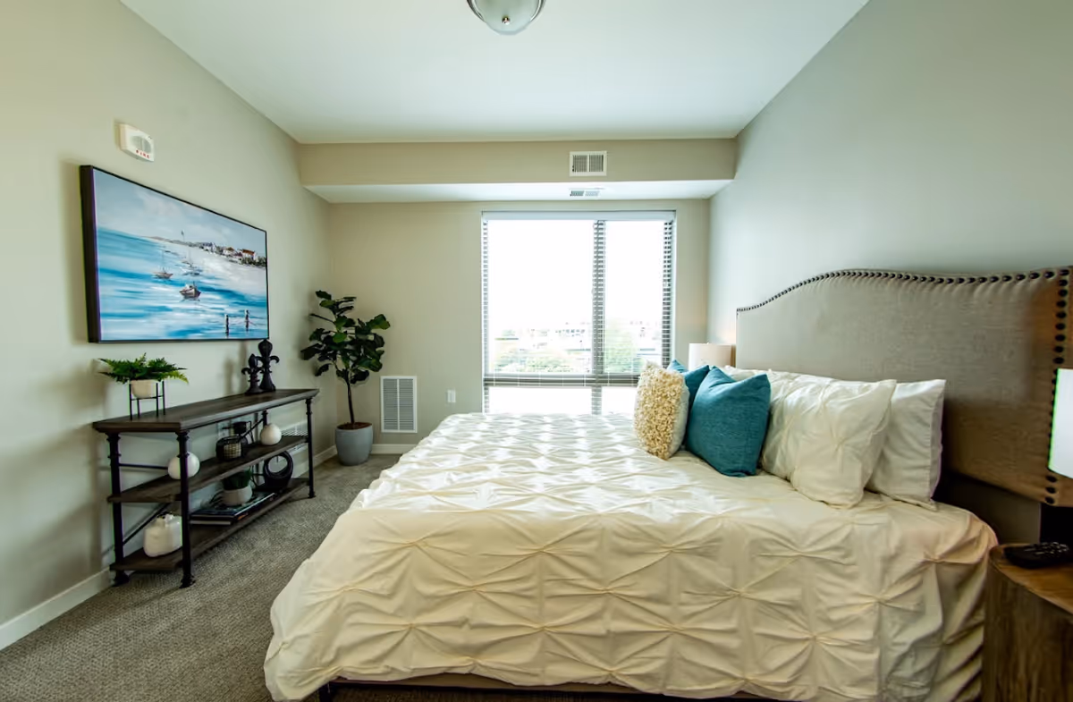 A bright bedroom with a large bed featuring a tufted beige headboard and white bedding accented with blue and beige pillows. There is a window with blinds letting in natural light, a wooden console table with decorative items, a potted plant, and a painting of a seaside scene on the wall.