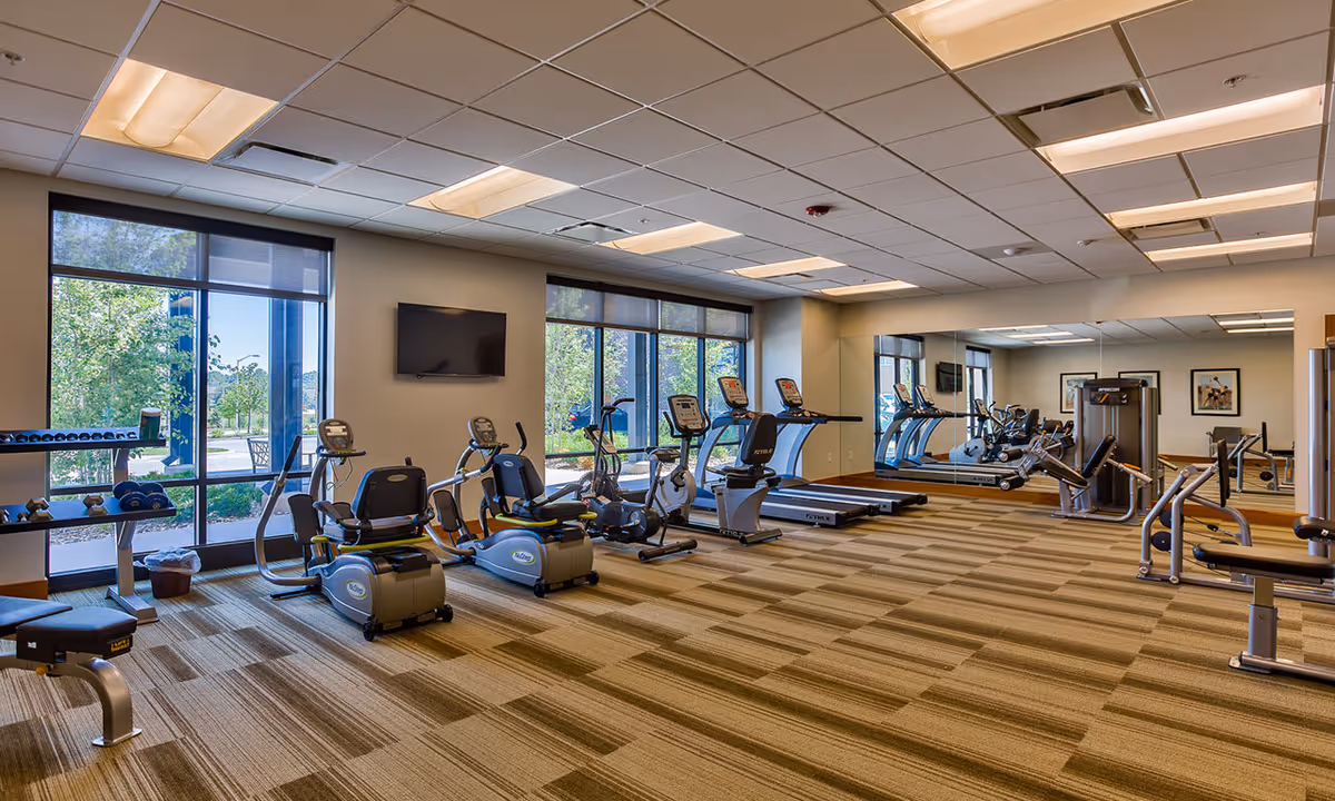 A spacious fitness room in a senior living facility with exercise equipment including recumbent bikes, treadmills, elliptical machines, weight benches, and dumbbells. Large windows allow natural light to fill the room, and a wall-mounted TV is visible. A large mirror covers one wall, reflecting the equipment and enhancing the sense of space.
