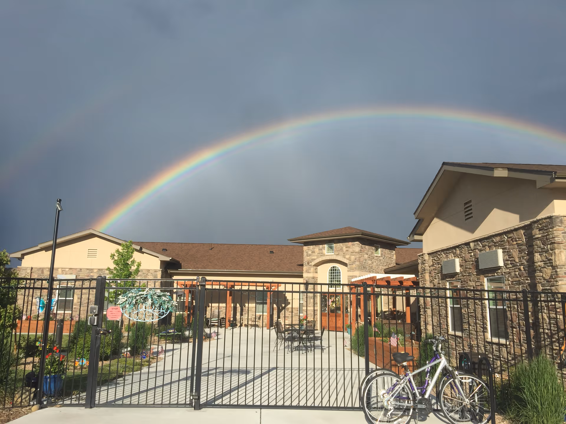 Gated exterior courtyard of a memory care building with stone façades and a double rainbow overhead.