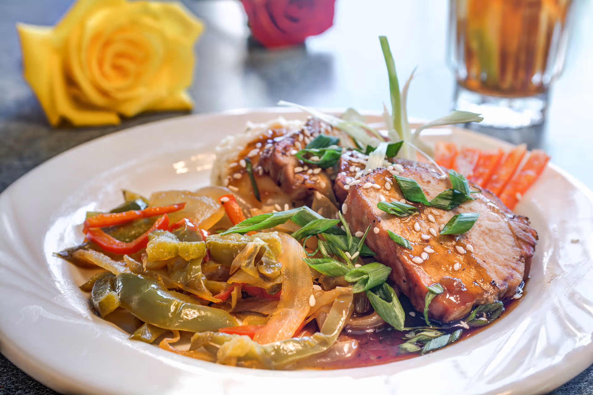 Close-up of a plated meal featuring glazed meat garnished with sesame seeds and green onions, sautéed peppers, and a drink and yellow rose in the background.