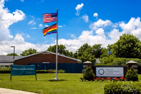Flags fly above a well-kept lawn and signs for The Trinity Community at Fairborn in front of a low brick building under a blue sky.
