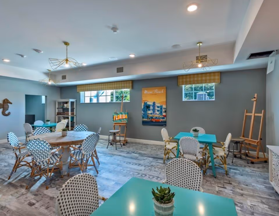 A bright and airy common room with several tables and chairs arranged for group seating. The walls are painted gray with two windows letting in natural light. There are decorative elements including a seahorse wall art, a Miami Beach poster, and an easel with a Florida sign. The floor has a patterned design, and the ceiling features modern light fixtures.