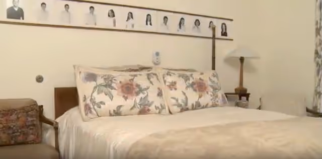 A bedroom with a bed featuring floral patterned pillows and a beige bedspread. There is a chair with a floral cushion on the left side and a wooden nightstand with a lamp on the right. Above the bed, there is a long framed photo collage of individual portraits.