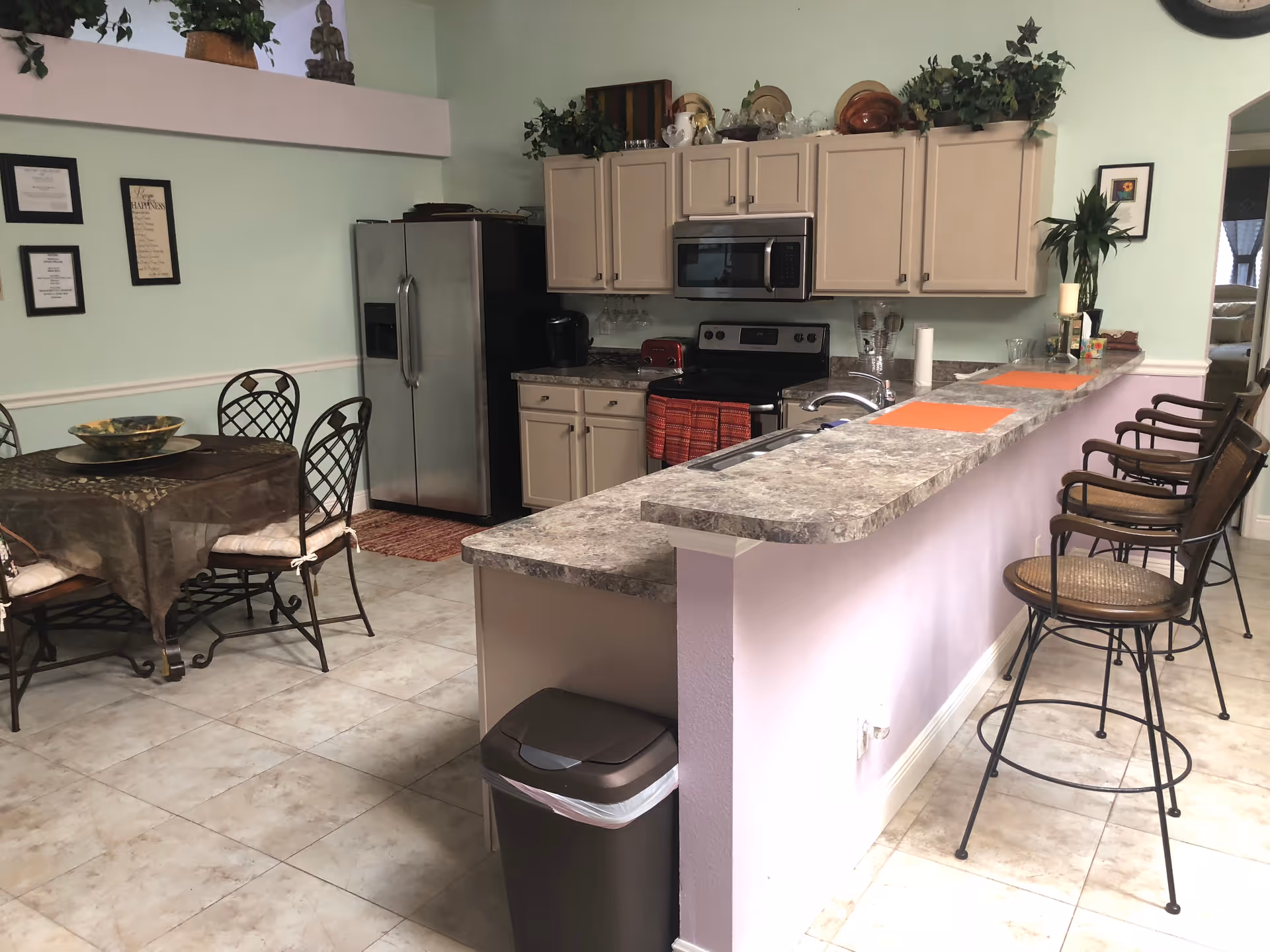 A kitchen and dining area in a senior living facility with light green walls and tiled floor. The kitchen features beige cabinets, a stainless steel refrigerator, stove, microwave, and a countertop with a sink and four bar stools. A round dining table with four chairs is set with a tablecloth and a decorative bowl. There are plants and decorative items on top of the cabinets and a few framed pictures on the walls.