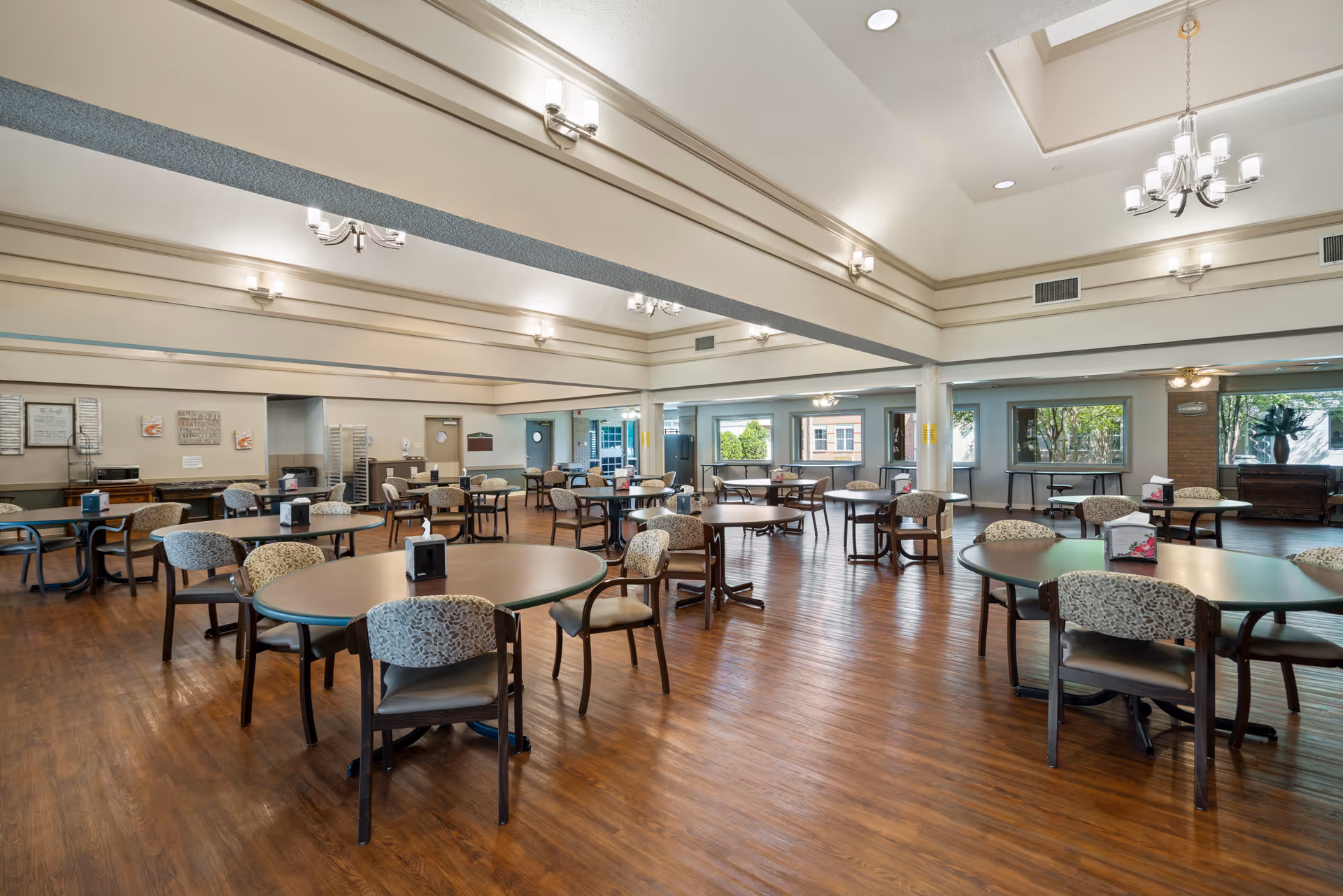 Spacious dining room with multiple round tables and cushioned chairs arranged neatly on a wooden floor. The room has large windows allowing natural light to enter, beige walls, and ceiling lights including chandeliers. The setting is clean and organized, suitable for group dining in a senior living facility.