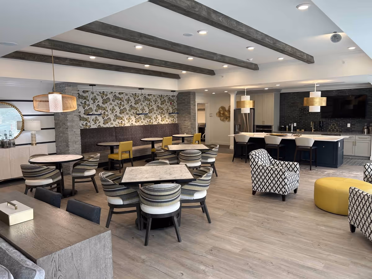 A spacious, modern common area in a senior living facility featuring multiple round tables with striped chairs, a kitchen island with bar stools, patterned armchairs, a yellow ottoman, and decorative ceiling beams. The walls have floral wallpaper and a large mirror, with pendant lighting above the kitchen island.