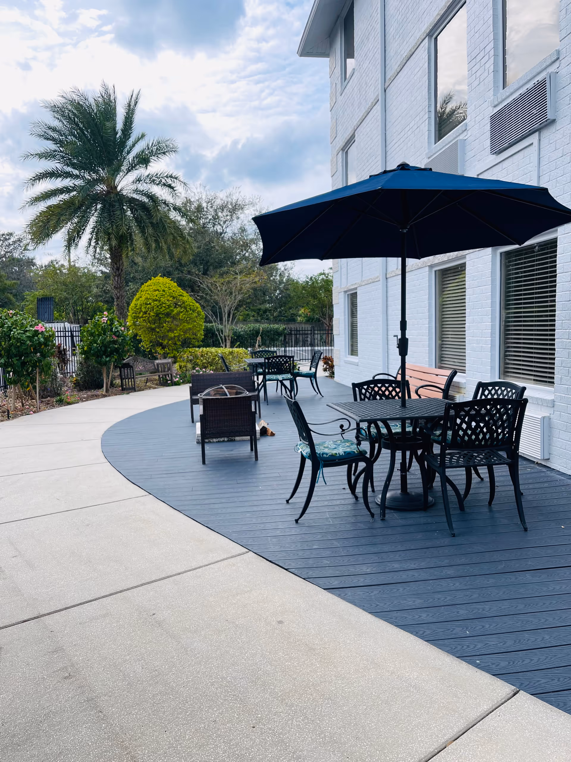 Outdoor patio area at Grace Manor Suites featuring a curved deck with black metal tables and chairs, some with cushions, and a large black umbrella. The patio is adjacent to a white brick building with several windows. Surrounding the patio are landscaped bushes, a palm tree, and other greenery under a partly cloudy sky.