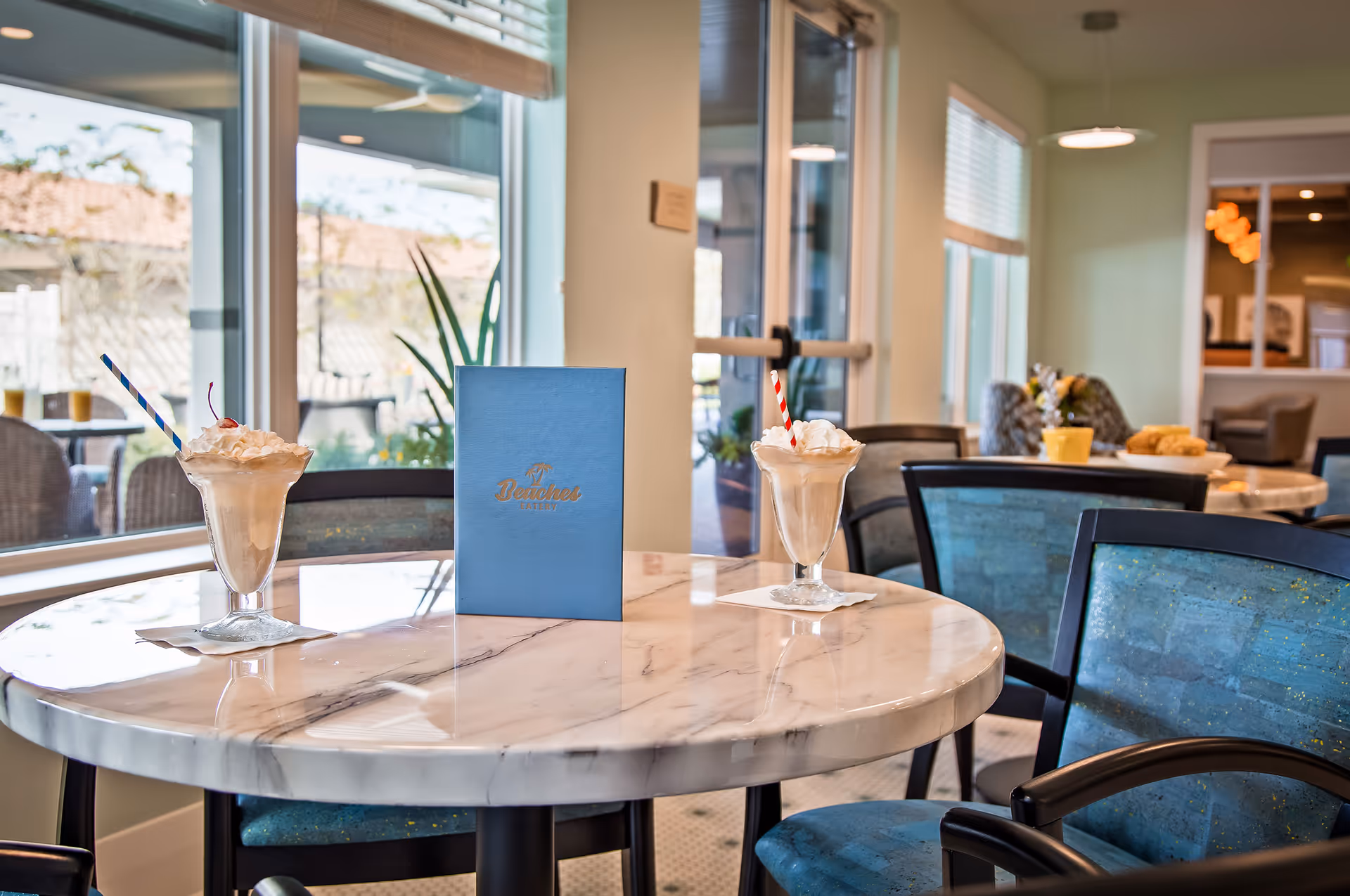 Round marble table with two milkshakes and a blue menu card in a bright dining area with upholstered chairs.