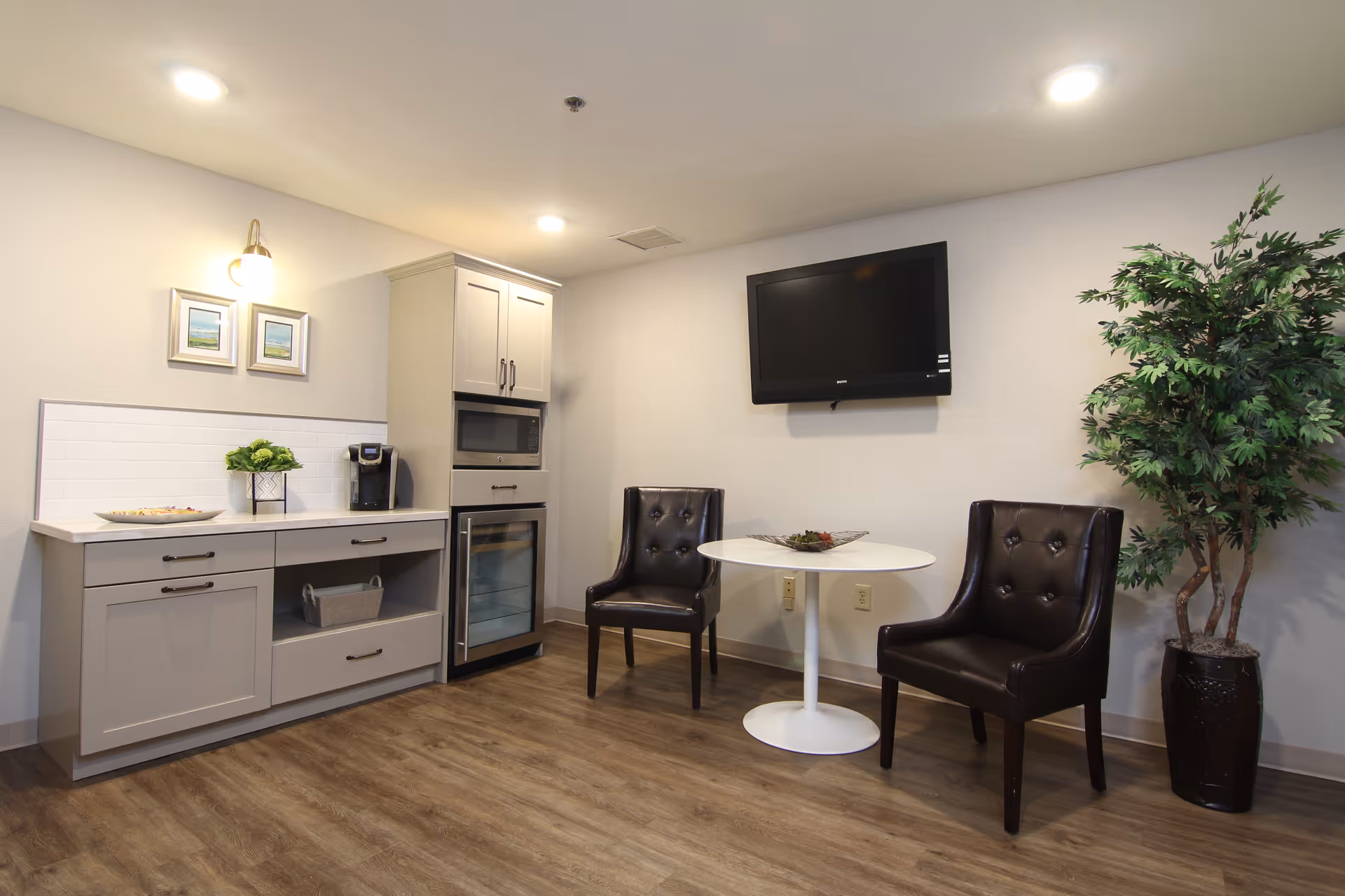 A small seating area in a nursing and rehab facility featuring two dark brown leather chairs around a white round table with a decorative centerpiece. Mounted on the wall above the table is a flat-screen TV. To the left, there is a kitchenette area with light gray cabinets, a countertop with a coffee maker, a small wine fridge, and two framed pictures on the wall. A tall potted plant is placed on the right side of the seating area. The floor is wood laminate and the walls are painted light gray.