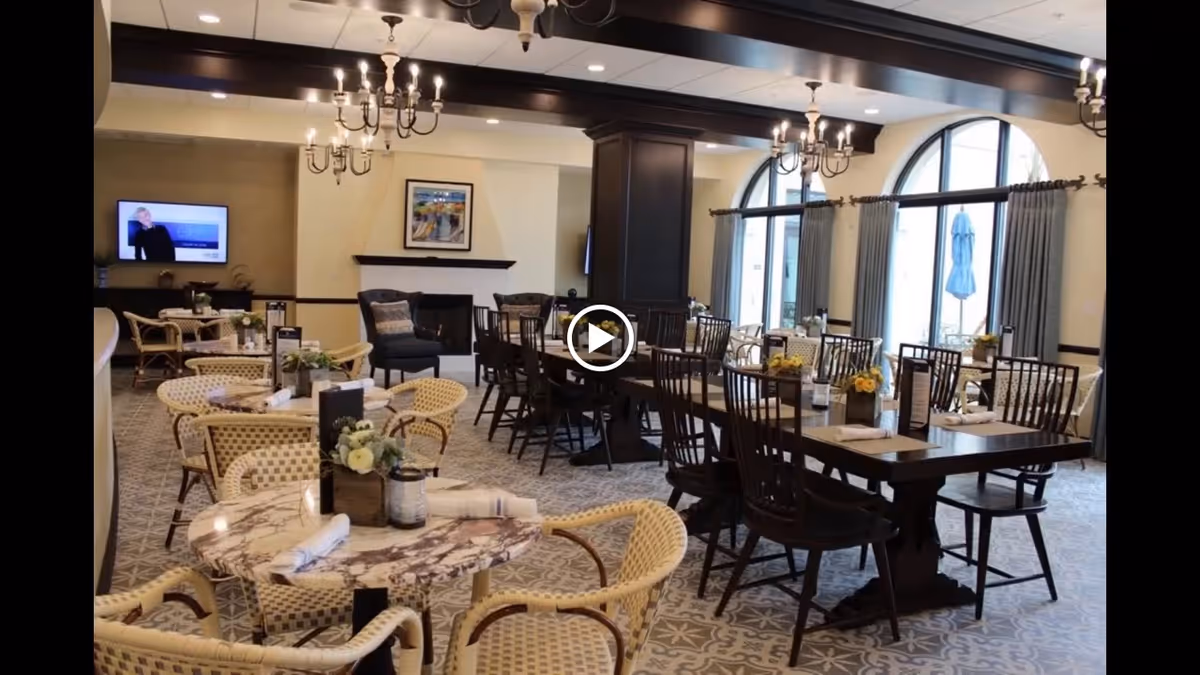 A spacious dining room with multiple tables and chairs arranged neatly. The room features large arched windows with curtains, chandeliers hanging from the ceiling, and a fireplace with a framed picture above it. Tables are set with napkins and small floral centerpieces.