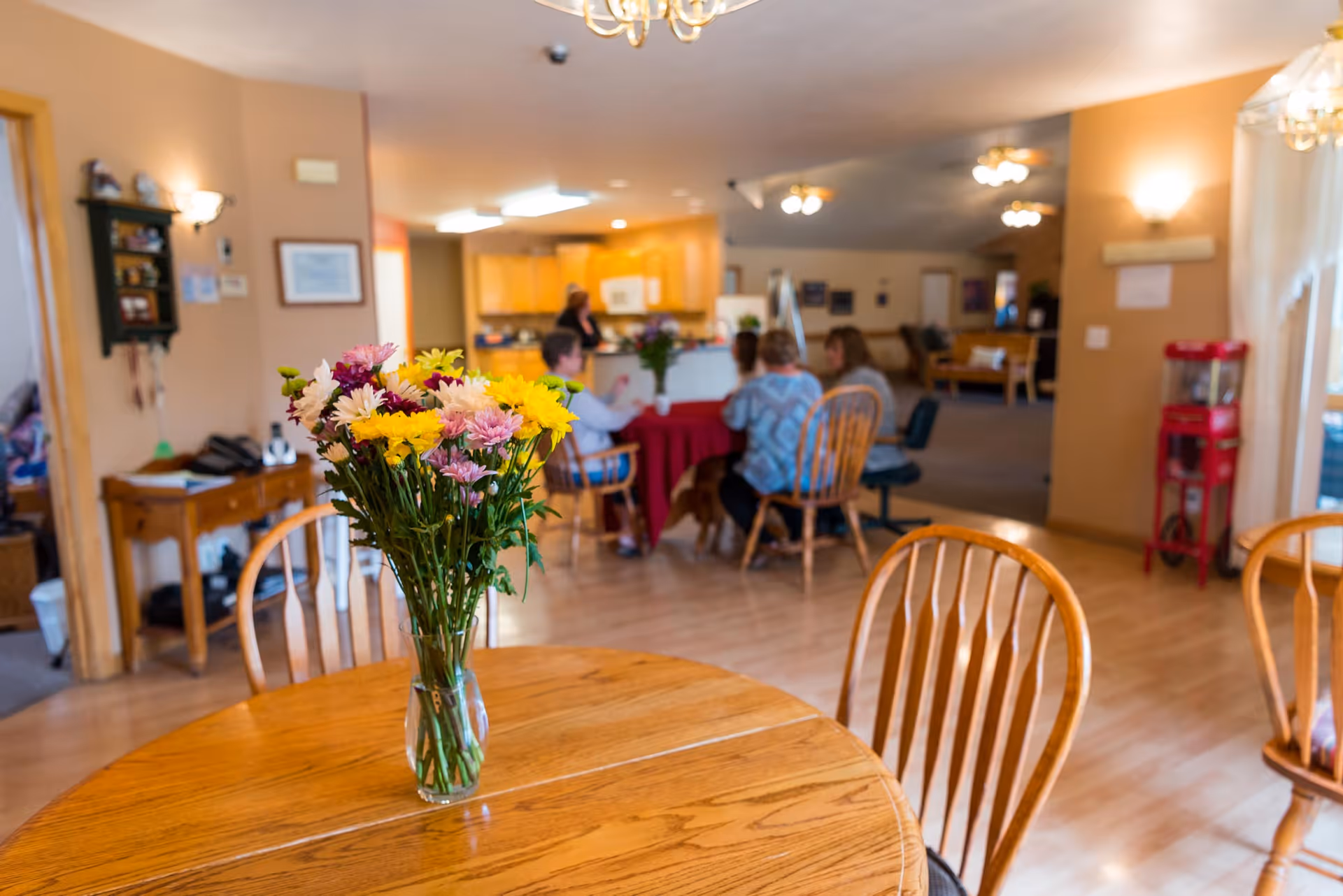 A cozy assisted living common area with a wooden round table in the foreground holding a vase of colorful flowers. In the background, several people are seated around another table, engaging in conversation. The room has warm lighting, wooden floors, and a kitchen area visible further back.