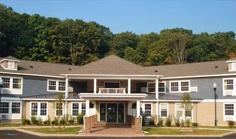 Front exterior view of a two-story senior living facility building with a covered entrance, multiple windows, and surrounded by greenery and trees under a clear blue sky.