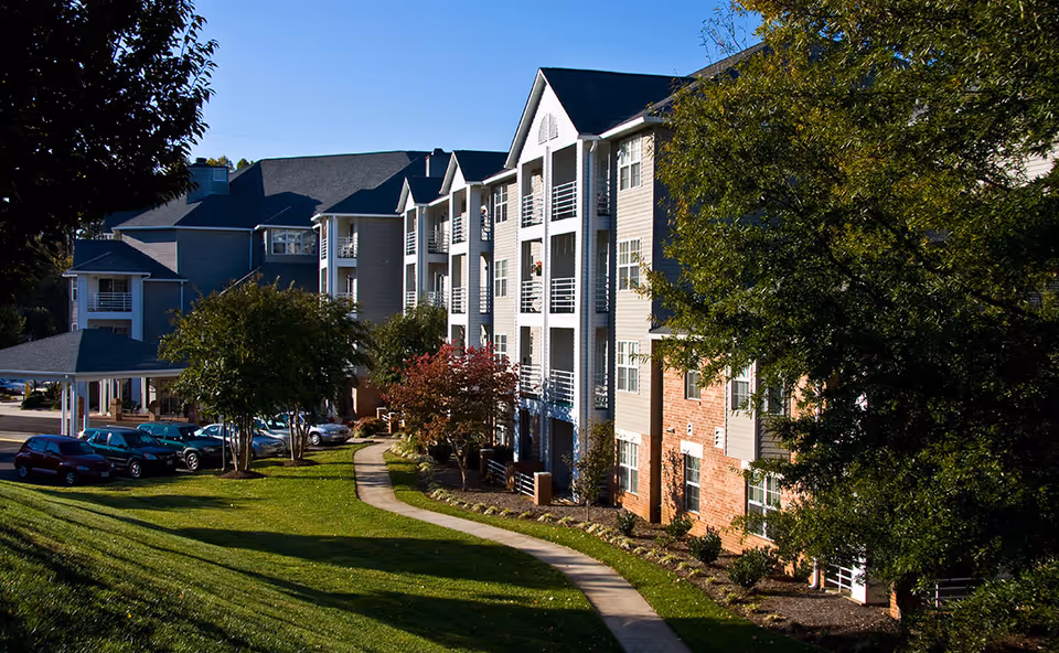 Exterior view of a multi-story retirement community building with balconies, surrounded by trees and a well-maintained lawn with a curved sidewalk. Several cars are parked near the building under a clear blue sky.