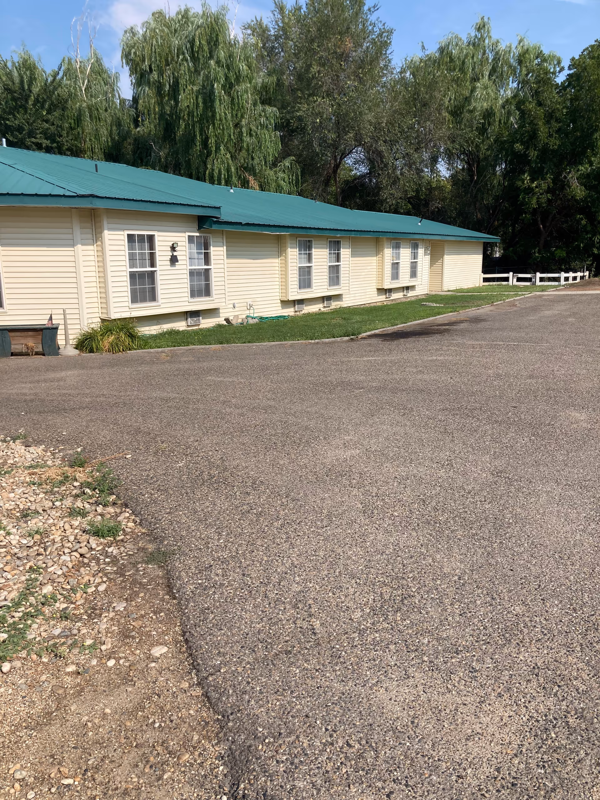 Exterior view of a single-story building with a green metal roof and beige siding, surrounded by trees and a paved driveway in front.
