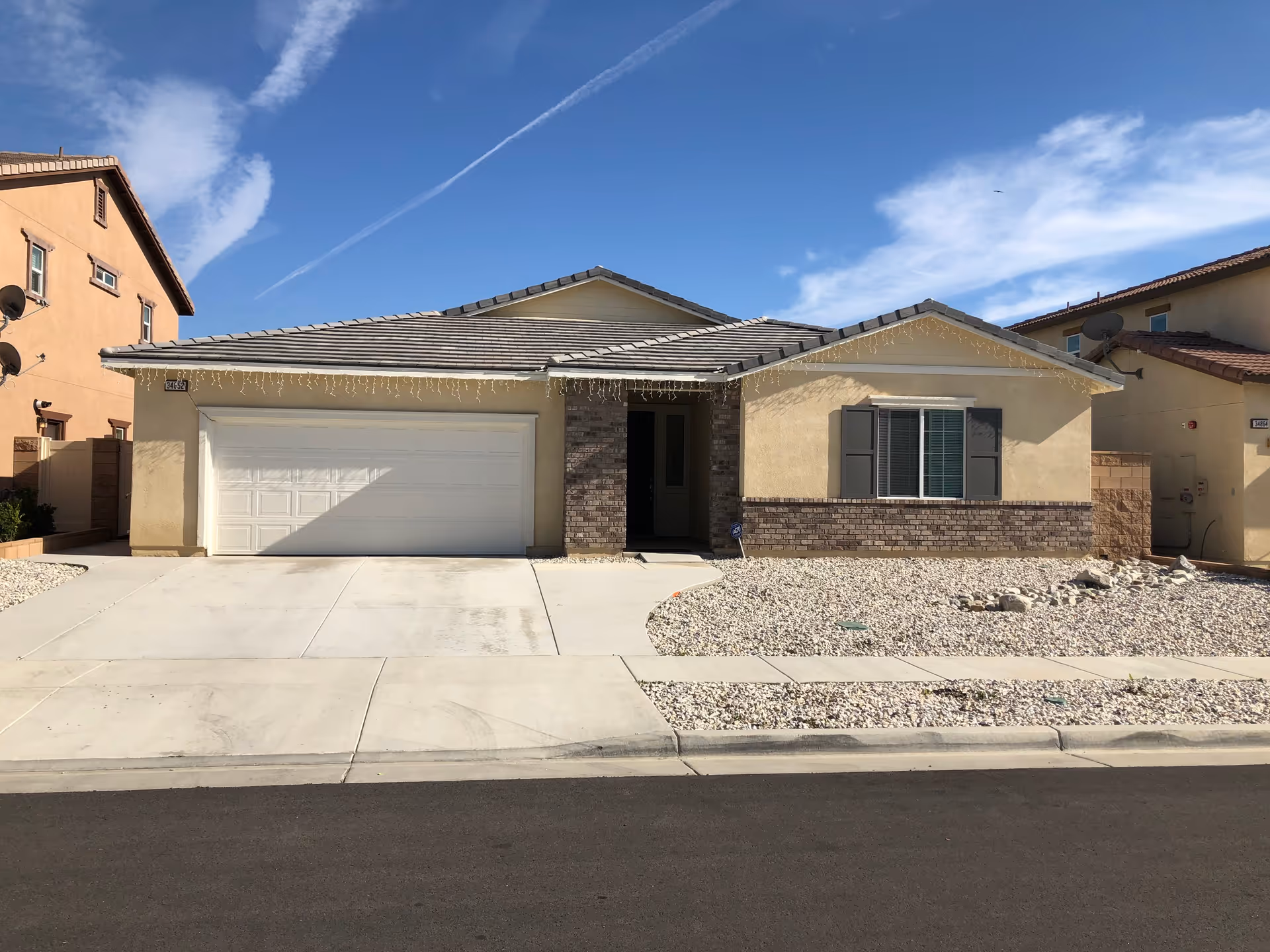 Single-story beige suburban house with a two-car garage, rock landscaping, and a clear blue sky.