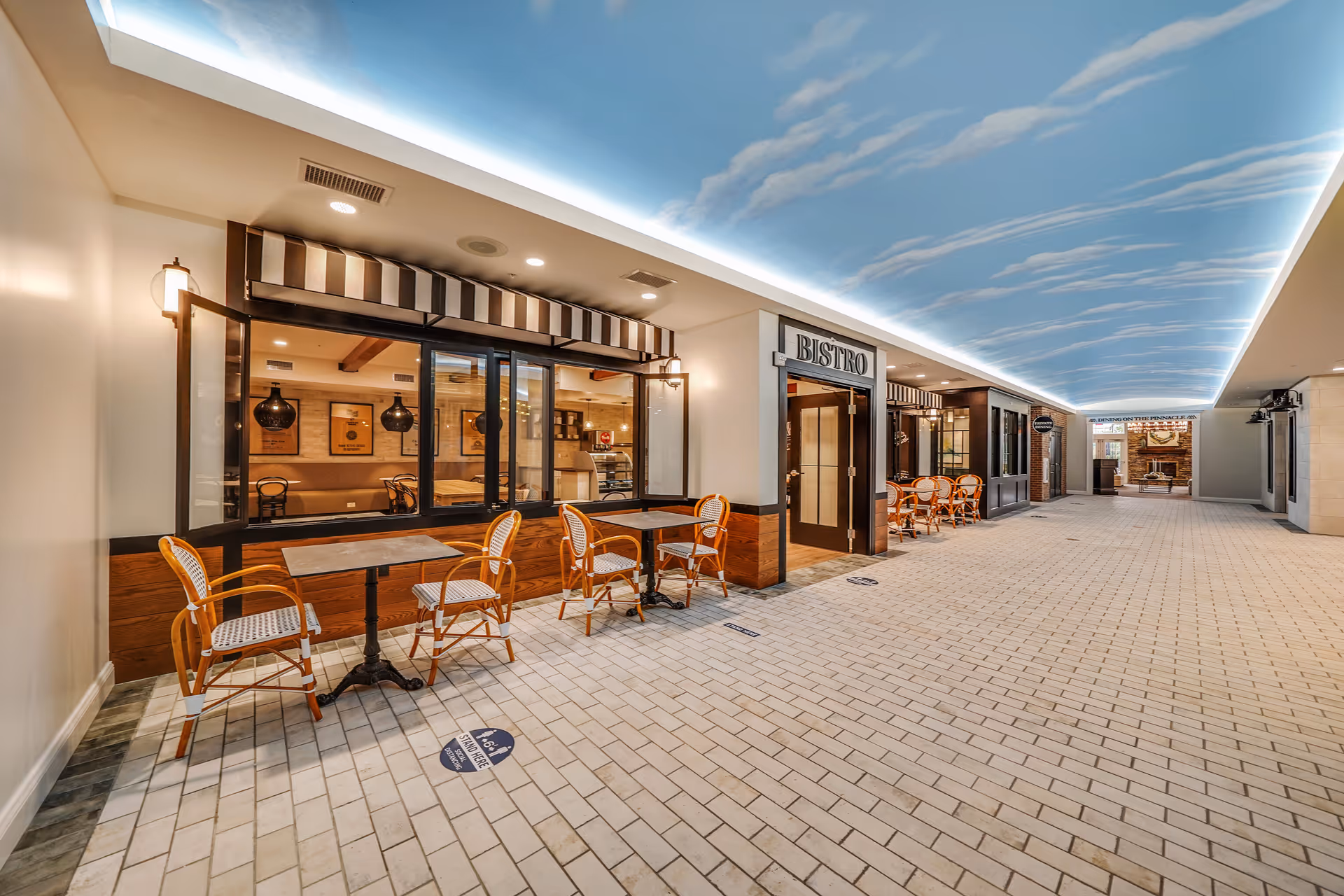 Indoor corridor featuring a Bistro storefront, wicker chairs, and a sky-painted ceiling over a tiled walkway.